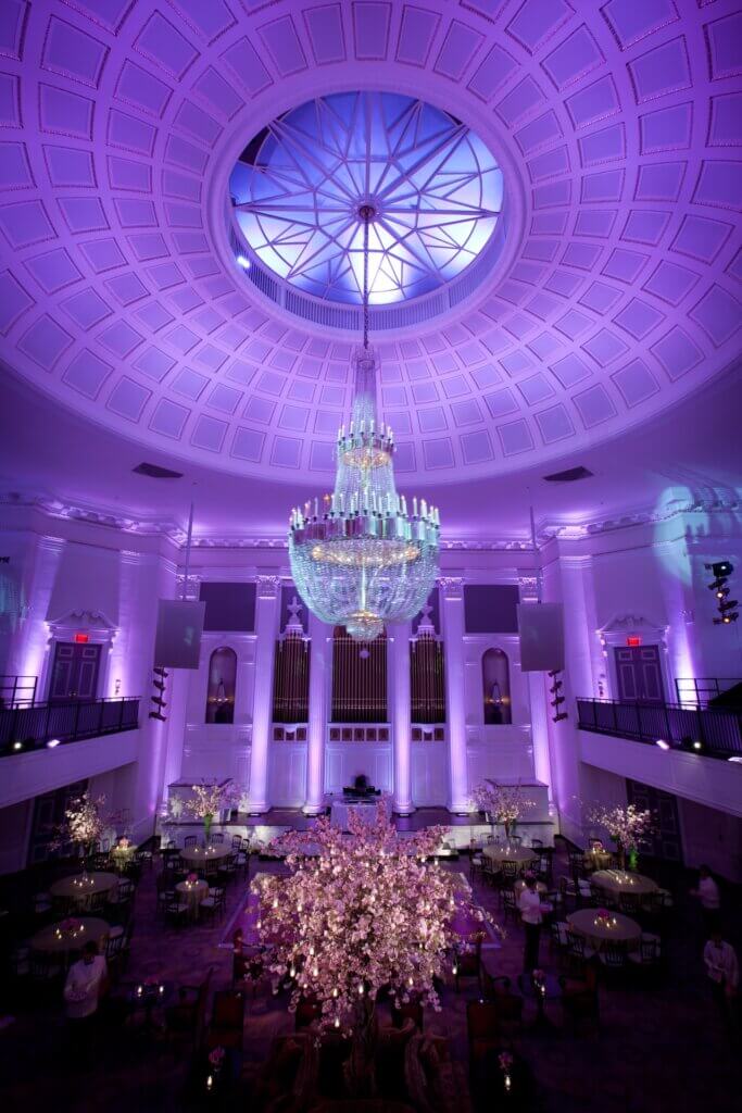 An elegant event space with a large domed ceiling and a grand chandelier. The room is bathed in purple lighting, with tall columns and decorative floral arrangements on tables below. The architectural details include ornate molding and a circular skylight.