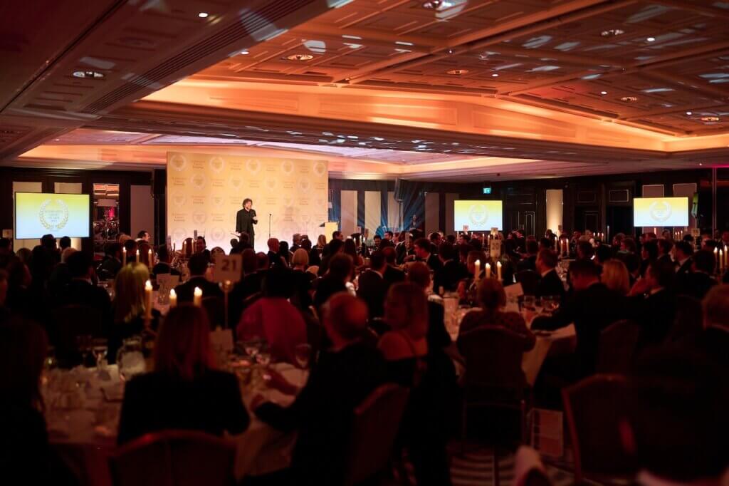 A formal awards event in a large banquet hall with round tables set for dinner. Guests in formal attire are seated, and a speaker is on stage in front of a branded backdrop. Two large screens display the event logo, and the room is warmly lit with orange tones.